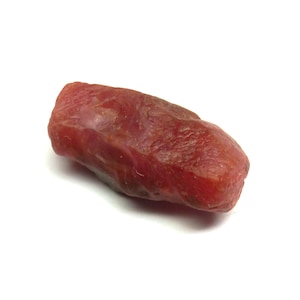 May include: A rough, reddish-brown gemstone with a textured surface. The elongated stone has a natural, organic shape and is set against a white background. The gemstone appears to be a raw, uncut mineral.