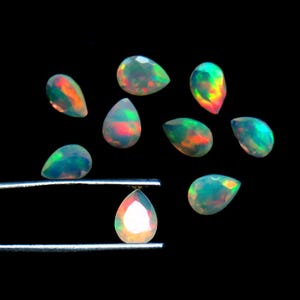 May include: A collection of pear-shaped opals with iridescent colors of red, orange, and green against a black background. One opal is held by tweezers. The opals vary in size and display a play of color.