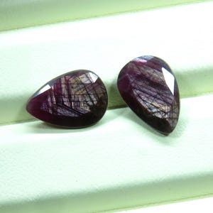 May include: Two pear-shaped, faceted gemstones with a deep burgundy color and internal striations. The gems have a polished, reflective surface, showcasing a mix of dark red and brown hues. The stones are likely rubies or similar precious stones.