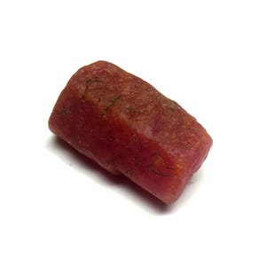 Puede incluir: Una gema de rubí en bruto y sin cortar, de color rojo intenso. El rubí tiene forma rectangular con una superficie ligeramente texturizada. La gema se muestra sobre un fondo blanco.