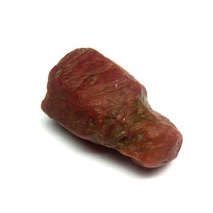 May include: A rough, reddish-brown gemstone with a textured surface. The stone has a natural, organic shape and appears to be unpolished. The color varies slightly, with some areas showing hints of green. The gemstone is set against a white background.