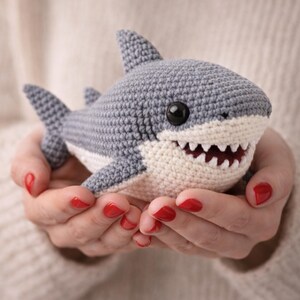 Könnte beinhalten: Handgehäkeltes Hai-Spielzeug in Grau und Weiß. Der Hai hat Flossen, ein Maul mit weißen Zähnen und schwarze Augen. Das Spielzeug ist weich und für Kinder konzipiert. Ein schönes Geschenk.