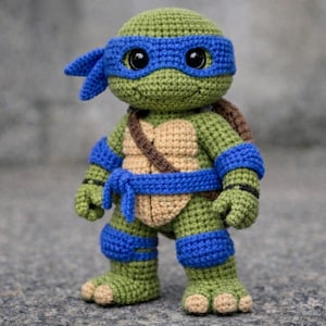 Puede incluir: Muñeco de Leonardo de las Tortugas Ninja hecho a ganchillo. El juguete hecho a mano presenta un cuerpo verde, una máscara azul y un caparazón marrón. El muñeco mide aproximadamente 20 cm de alto y está sobre una superficie gris.