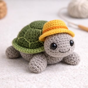 Puede incluir: Una tortuga tejida a mano con caparazón verde y cuerpo gris. La tortuga lleva un sombrero amarillo con una banda naranja. Tiene ojos negros y una cara sonriente. Juguete artesanal.