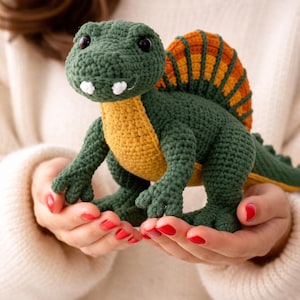 May include: A handmade crochet dinosaur toy in shades of green, yellow, and orange. The dinosaur has a distinctive sail on its back and is held in someone's hands. The toy is a cute and cuddly representation of a prehistoric creature.