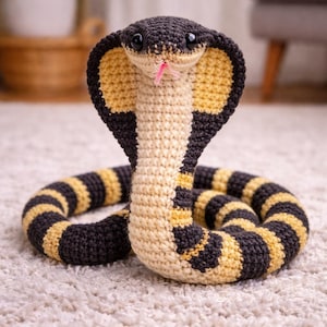 Könnte beinhalten: Ein gehäkeltes Kobra-Schlangenspielzeug in Schwarz, Beige und Gelb. Die Schlange hat eine ausgestellte Kapuze, einen beigen Bauch und eine rosa Zunge. Der Körper ist mit Schwarz und Gelb gestreift. Das Spielzeug liegt auf einem weißen Teppich.