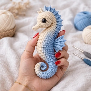 Puede incluir: Un caballito de mar de ganchillo hecho a mano en crema y azul. El caballito de mar tiene un diseño detallado con una cola rizada y aletas. Se sostiene en una mano, con hilo y ganchillos en el fondo. El caballito de mar mide aproximadamente 20 cm de largo.