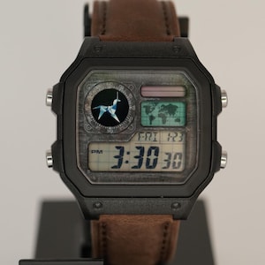 May include: Black digital watch with a brown leather strap. The watch face shows the time as 3:30:30 PM, a world map, and a compass design. The watch is on a black display stand with the Casio logo.