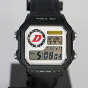 May include: Black digital watch with a white face, showing the time as 5:08:03 PM. The watch has a black strap and is labelled "WORLD TIME" and "ILLUMINATOR". The watch face features a "D" logo and the Casio brand.