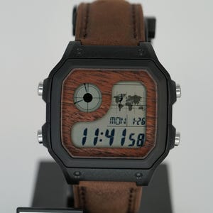 Pode incluir: Relógio digital preto com pulseira de couro castanho e mostrador com efeito de madeira. O relógio exibe a hora, a data e um gráfico do mapa-múndi. O relógio está em um suporte preto com os logotipos "KW Customs" e "Casio".