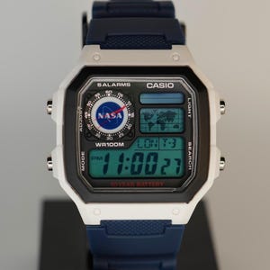 Puede incluir: Un reloj digital Casio con correa blanca y azul marino. La esfera del reloj muestra la hora, la fecha y un logotipo de la NASA. El reloj también tiene el texto "10 YEAR BATTERY". El reloj se muestra en un soporte negro.