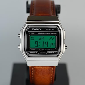 May include: A Casio F-91W digital watch with a silver case and a brown leather strap. The watch face displays the time, date, and day of the week in green digits. The watch is displayed on a black stand with the Casio logo.