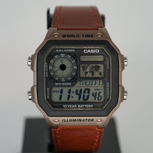 May include: A Casio digital watch with a brown leather strap. The watch face is black with a digital display showing the time. The watch has a bronze-colored case and features the text "CASIO" and "ILLUMINATOR". The watch is displayed on a black stand.