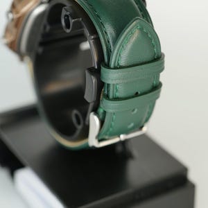 May include: Close-up of a wristwatch with a green leather band and a silver buckle. The watch face is partially visible, with a gold-colored bezel. The watch is displayed on a black stand against a white background.