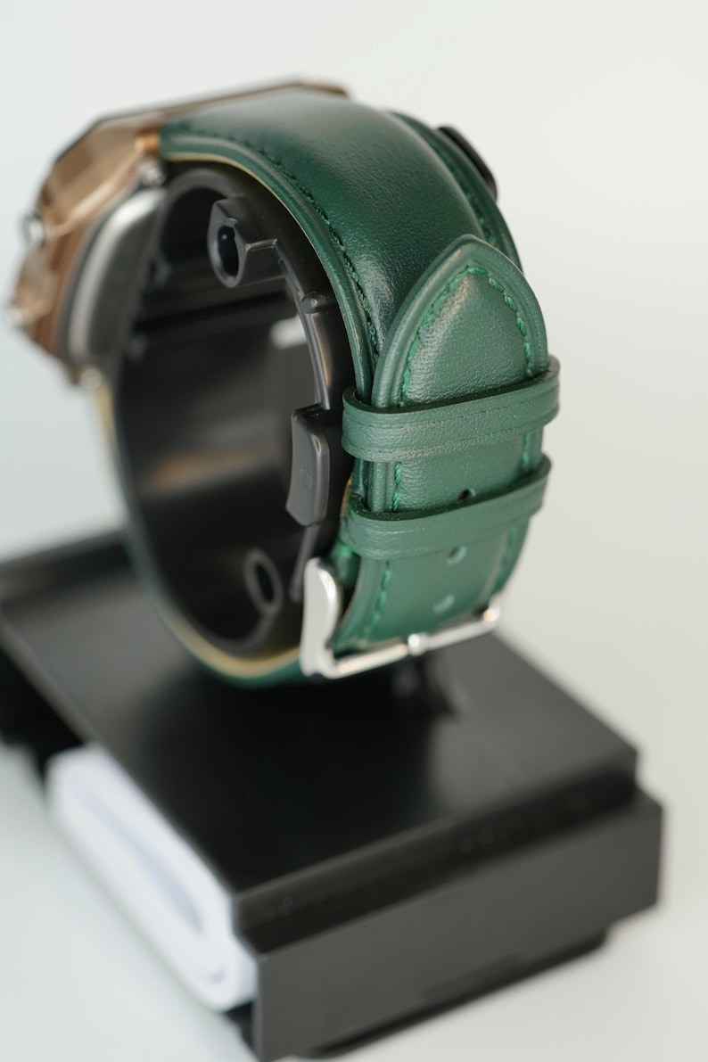 May include: Close-up of a wristwatch with a green leather band and a silver buckle. The watch face is partially visible, with a gold-colored bezel. The watch is displayed on a black stand against a white background.