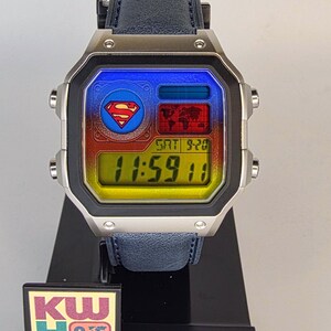 May include: A silver-toned Casio digital watch with a blue leather band. The watch face features the Superman logo and a colorful display. The time reads 11:59:11. The watch is displayed on a black stand with a "KW Customs" logo.