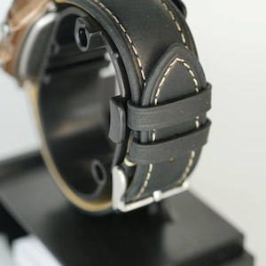 May include: Close-up of a black leather watch band with white stitching. The watch band is attached to a watch with a bronze-colored face. The watch is displayed on a black stand.
