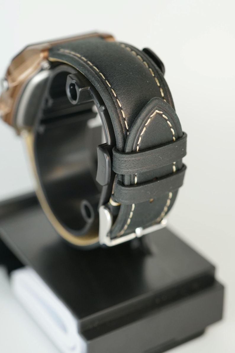 May include: Close-up of a black leather watch band with white stitching. The watch band is attached to a watch with a bronze-colored face. The watch is displayed on a black stand.