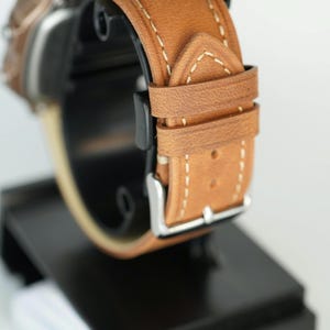May include: A brown leather watch band with white stitching and a silver buckle. The watch band is displayed on a black stand, showcasing its design and craftsmanship. The band's color is a warm, light brown.