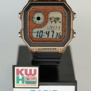 May include: A digital watch with a black band and a gold-colored case. The watch face displays the time, a world map, and the text "WORLD TIME" and "ILLUMINATOR". The watch is on a black display stand with the text "KW HO" and "CASIO".