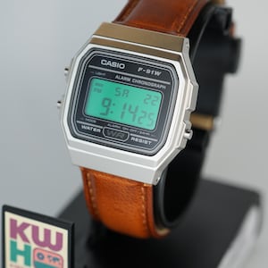 Puede incluir: Un reloj digital Casio F-91W con caja plateada y correa de cuero marr&oacute;n. La esfera muestra la hora, el d&iacute;a y la fecha. El reloj est&aacute; sobre un expositor negro. El texto "CASIO" es visible.