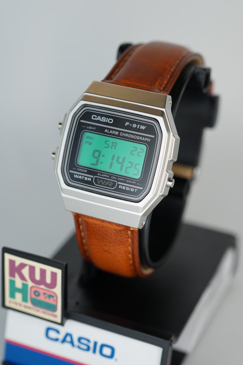 Puede incluir: Un reloj digital Casio F-91W con caja plateada y correa de cuero marr&oacute;n. La esfera muestra la hora, el d&iacute;a y la fecha. El reloj est&aacute; sobre un expositor negro. El texto "CASIO" es visible.
