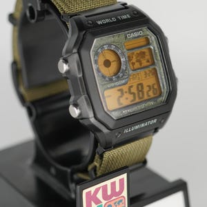 Puede incluir: Reloj digital Casio negro con correa de nailon verde. La esfera muestra la hora, la fecha y las funciones de hora mundial. El reloj est&aacute; sobre un expositor negro con el texto "CASIO" y "KW Watch House".