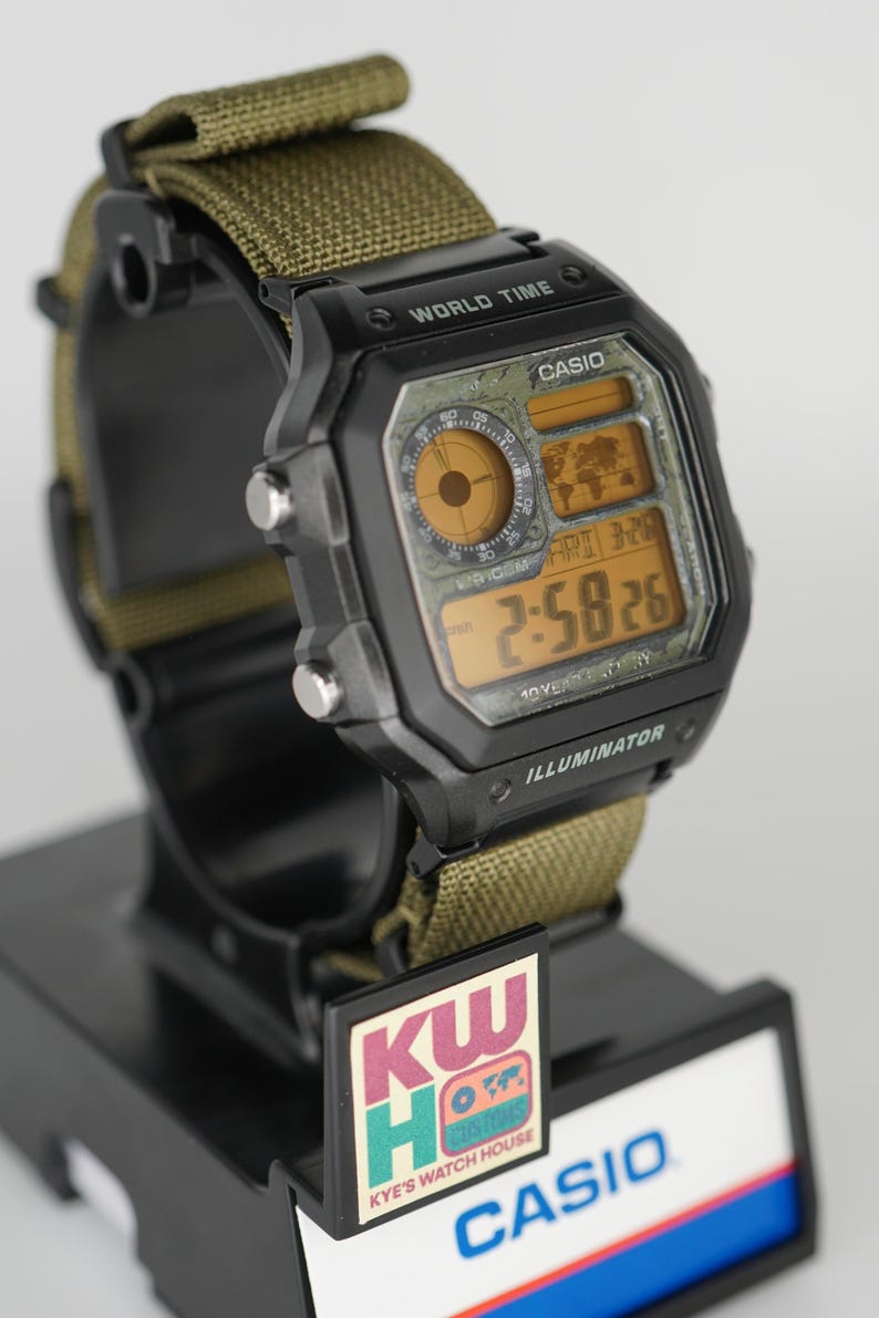 Puede incluir: Reloj digital Casio negro con correa de nailon verde. La esfera muestra la hora, la fecha y las funciones de hora mundial. El reloj est&aacute; sobre un expositor negro con el texto "CASIO" y "KW Watch House".