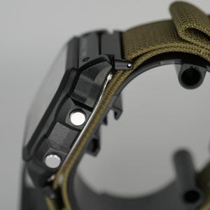 Puede incluir: Primer plano de un reloj digital negro con una correa de nailon verde. El reloj tiene dos botones plateados en el lateral y una esfera rectangular. La correa es de color verde oliva.