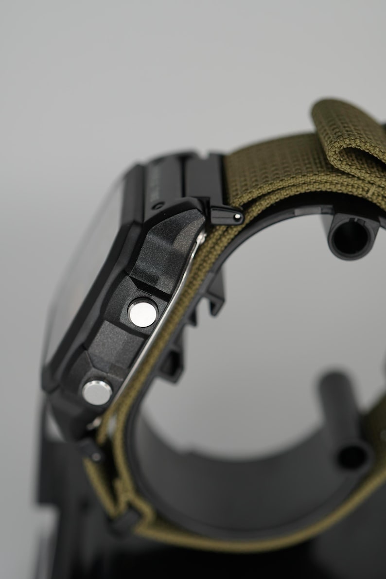 Puede incluir: Primer plano de un reloj digital negro con una correa de nailon verde. El reloj tiene dos botones plateados en el lateral y una esfera rectangular. La correa es de color verde oliva.