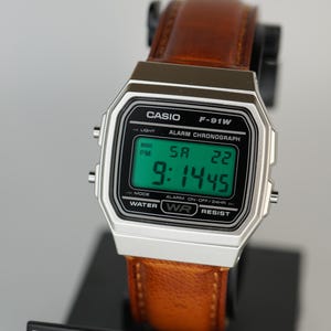 Puede incluir: Un reloj digital Casio F-91W con caja plateada y correa de cuero marr&oacute;n. El reloj muestra la hora, el d&iacute;a y la fecha en una pantalla digital verde. El reloj es resistente al agua.