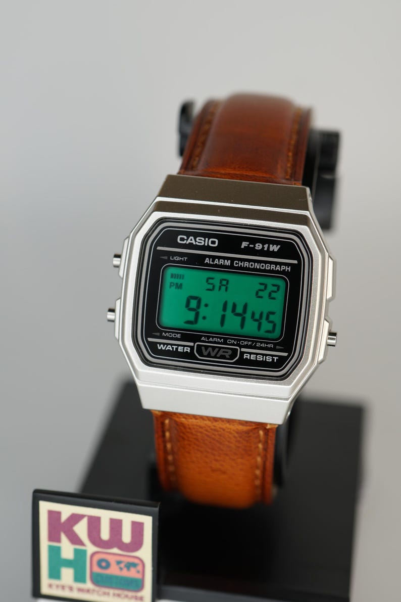 Puede incluir: Un reloj digital Casio F-91W con caja plateada y correa de cuero marr&oacute;n. El reloj muestra la hora, el d&iacute;a y la fecha en una pantalla digital verde. El reloj es resistente al agua.
