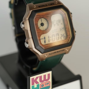 May include: A Casio digital watch with a wood-grain face and a green leather strap. The watch displays the time and world time functions. The watch is on a black display stand with the text "WORLD TIME" and "ILLUMINATOR".