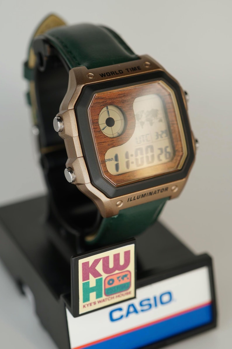 May include: A Casio digital watch with a wood-grain face and a green leather strap. The watch displays the time and world time functions. The watch is on a black display stand with the text "WORLD TIME" and "ILLUMINATOR".