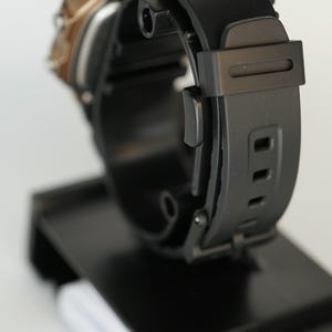 May include: A black digital watch with a black rubber strap is displayed on a black stand. The watch face has a gold-colored trim. The strap has multiple holes for adjustment. The watch is designed for everyday wear.