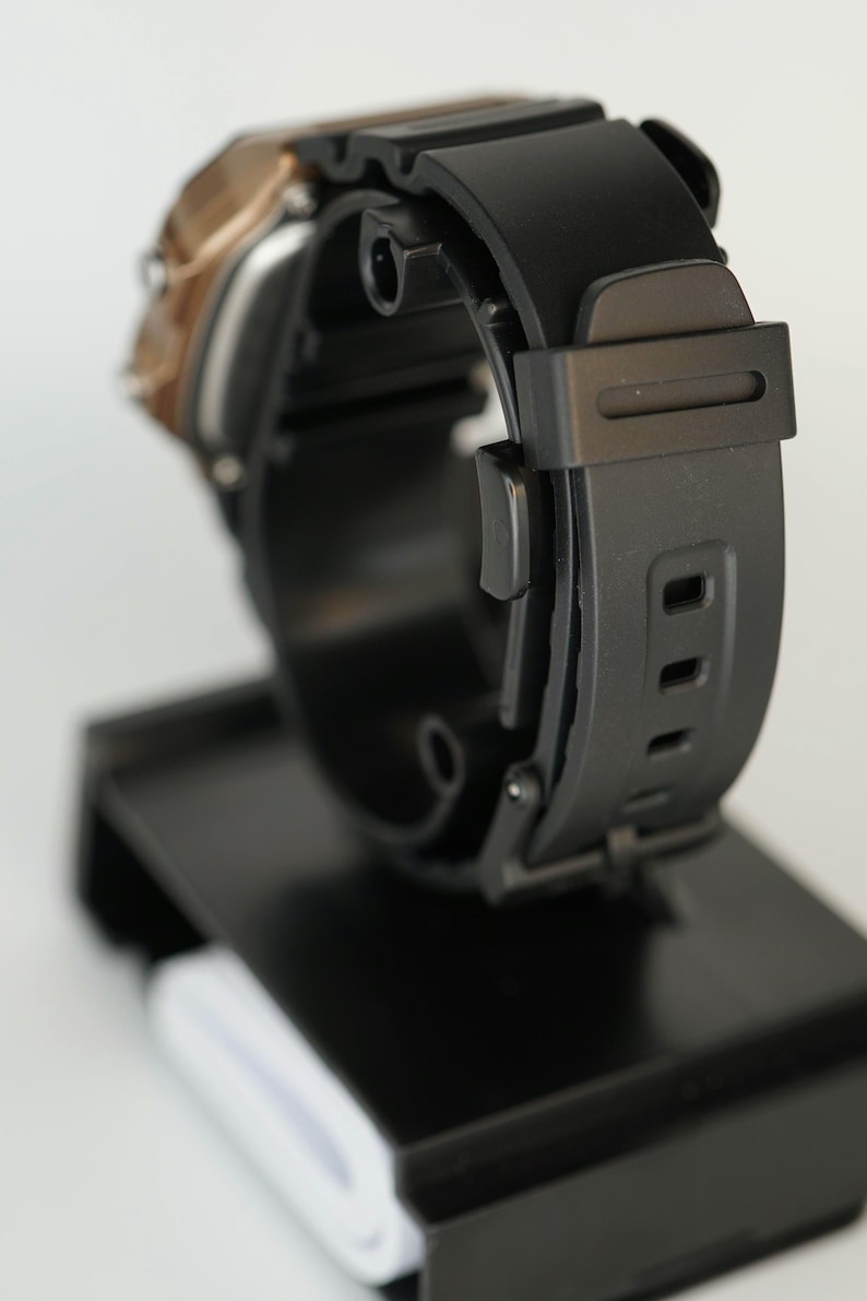 May include: A black digital watch with a black rubber strap is displayed on a black stand. The watch face has a gold-colored trim. The strap has multiple holes for adjustment. The watch is designed for everyday wear.