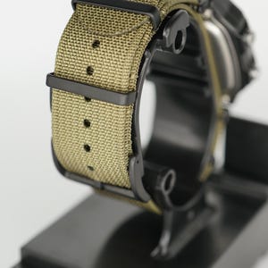 Puede incluir: Primer plano de un reloj de pulsera con correa de lona verde oliva y herrajes negros. El reloj se muestra sobre un soporte negro, mostrando la textura de la correa y la esfera oscura. La correa mide aproximadamente 2,5 cm de ancho.