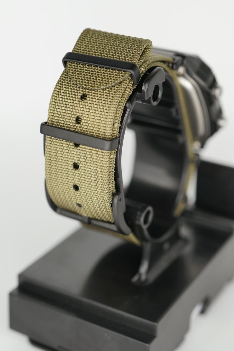 Puede incluir: Primer plano de un reloj de pulsera con correa de lona verde oliva y herrajes negros. El reloj se muestra sobre un soporte negro, mostrando la textura de la correa y la esfera oscura. La correa mide aproximadamente 2,5 cm de ancho.