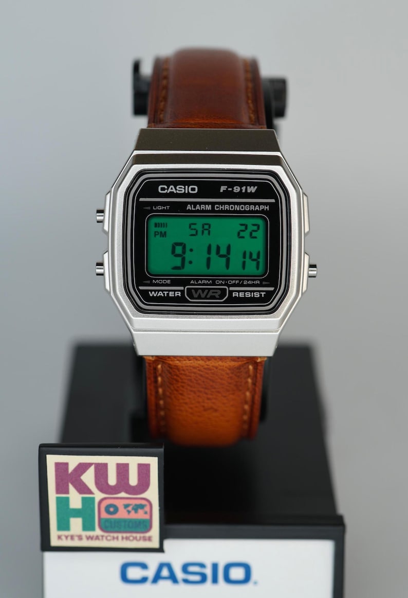 Puede incluir: Un reloj digital Casio F-91W con caja plateada y correa de cuero marr&oacute;n. La esfera muestra la hora, la fecha y el d&iacute;a de la semana en d&iacute;gitos verdes. El reloj se exhibe sobre un soporte negro con el logotipo de Casio.