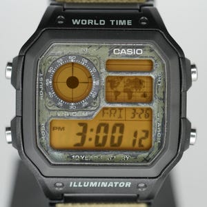 Puede incluir: Reloj digital Casio negro con esfera verde estilo camuflaje. El reloj muestra la hora a las 3:00 PM y la fecha Vie 3-26. El reloj tiene el texto "WORLD TIME" e "ILLUMINATOR" en la esfera.