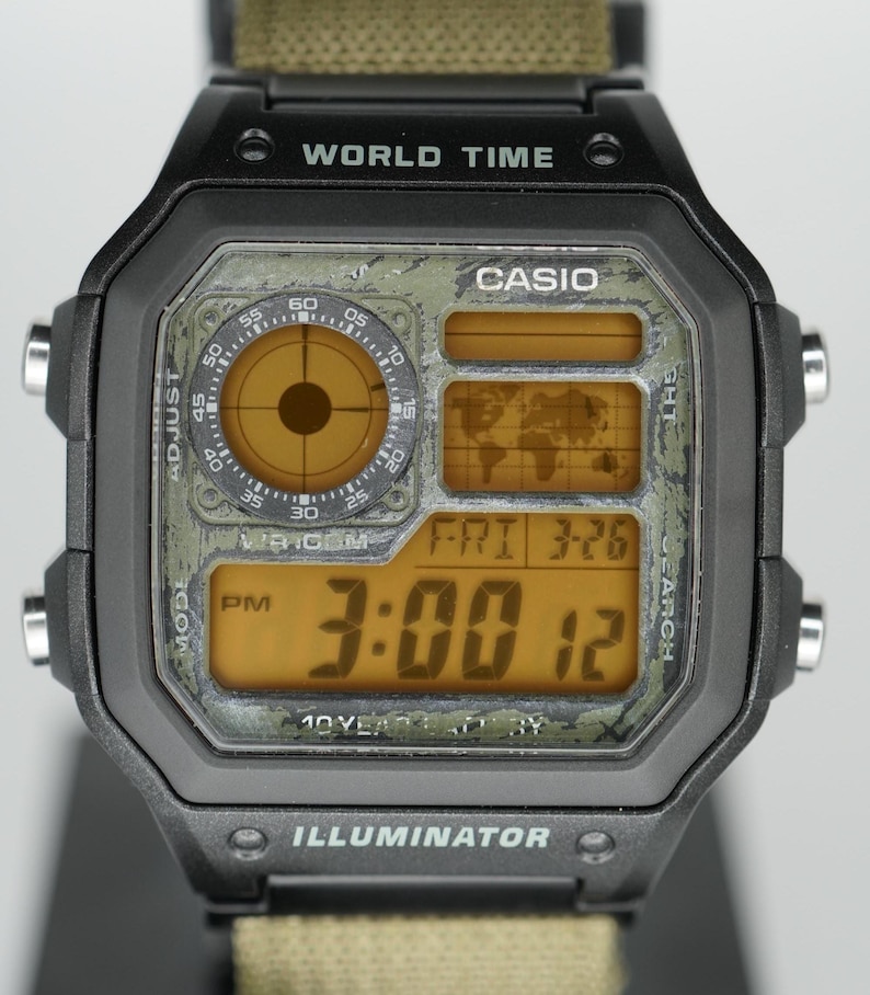 Puede incluir: Reloj digital Casio negro con esfera verde estilo camuflaje. El reloj muestra la hora a las 3:00 PM y la fecha Vie 3-26. El reloj tiene el texto "WORLD TIME" e "ILLUMINATOR" en la esfera.