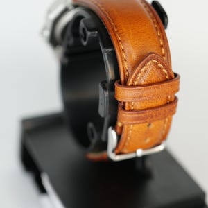 Puede incluir: Primer plano de un reloj de pulsera con una correa de cuero marr&oacute;n. La correa est&aacute; cosida con un hilo m&aacute;s claro y tiene una hebilla plateada. El reloj se muestra sobre un soporte negro sobre un fondo blanco.