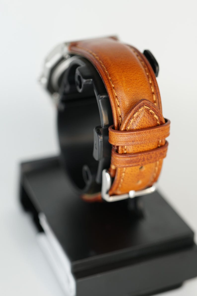 Puede incluir: Primer plano de un reloj de pulsera con una correa de cuero marr&oacute;n. La correa est&aacute; cosida con un hilo m&aacute;s claro y tiene una hebilla plateada. El reloj se muestra sobre un soporte negro sobre un fondo blanco.