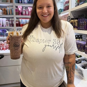 Pode incluir: Uma mulher vestindo uma camiseta branca com a frase "Chapter 39 yourself" em escrita preta, segurando um café gelado da Gloria Jean's. O fundo mostra prateleiras repletas de produtos de beleza.