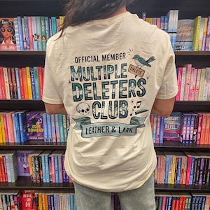 Puede incluir: Camiseta color crema con el texto "OFFICIAL MEMBER MULTIPLE DELETERS CLUB LEATHER & LARK". El diseño incluye una calavera, un pájaro y notas musicales. La camiseta se lleva delante de una estantería.
