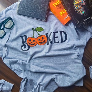 Puede incluir: Una camiseta de manga larga gris jaspeado con la palabra "BOOKED" en negro, con dos cerezas de calabaza naranja. La camiseta se combina con unas gafas de montura verde y varios libros.
