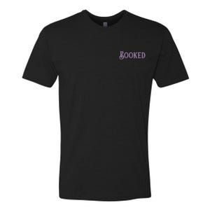 Puede incluir: Camiseta negra con la palabra "BOOKED" en escritura lila claro en el pecho. La camiseta tiene mangas cortas y cuello redondo. La tela parece ser un material suave y c&oacute;modo.