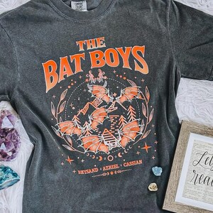 Puede incluir: Camiseta gris oscuro con el texto "THE BAT BOYS" en naranja. La camiseta presenta un gráfico naranja de murciélagos, árboles y estrellas. Los nombres Rhysand, Azriel y Cassian están impresos debajo.