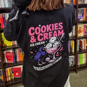 Puede incluir: Camiseta negra con un gr&aacute;fico rosa y blanco que dice "Cookies & Cream Ice Cream". El dise&ntilde;o incluye un cuervo, un helado y el texto "Tastes better because it's made right with more cream than the".