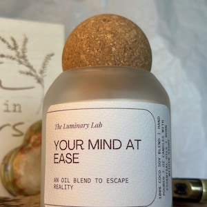 May include: A frosted glass candle jar with a cork lid. The label reads "YOUR MIND AT EASE" and "AN OIL BLEND TO ESCAPE REALITY". The candle is a 100% coco soy blend with essential oils. The Luminary Lab.
